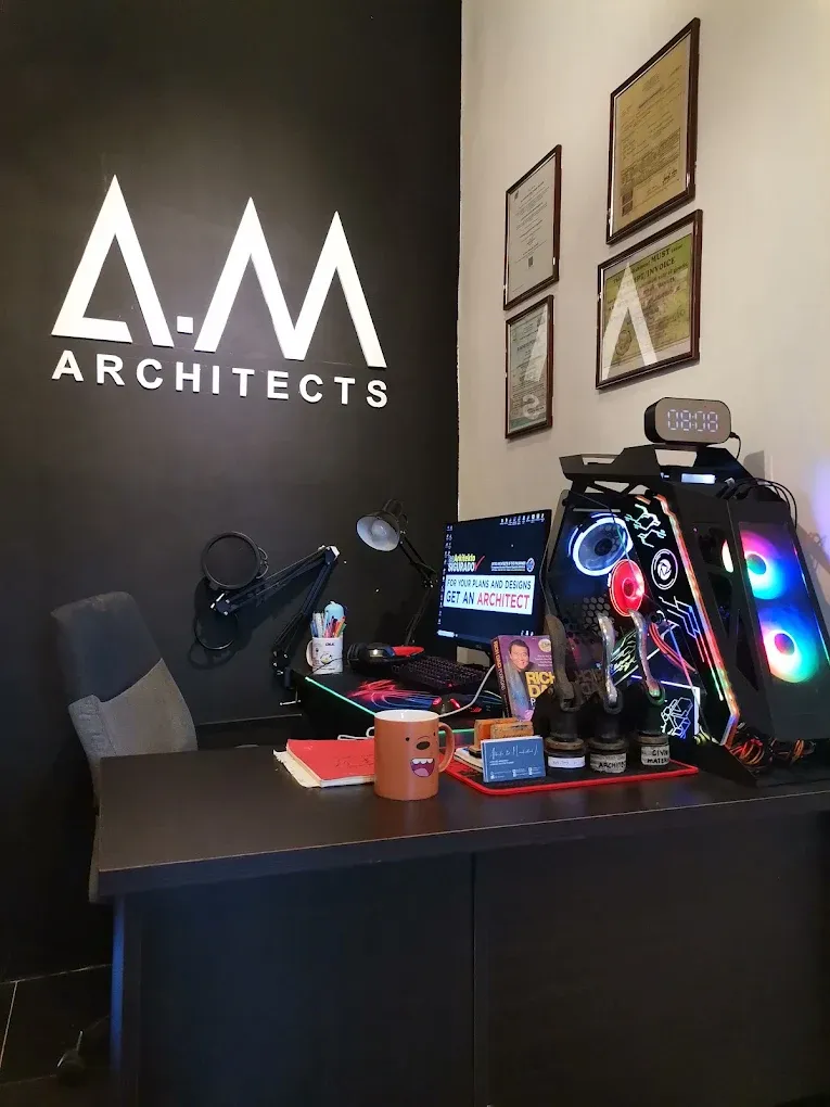 A desk beneath the white A.M Architects logo on a black wall, with framed certificates and a lit-up computer