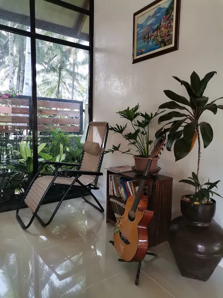 A lounge corner with a reclining chair, a guitar and indoor plants beside a glass wall open to the palm trees