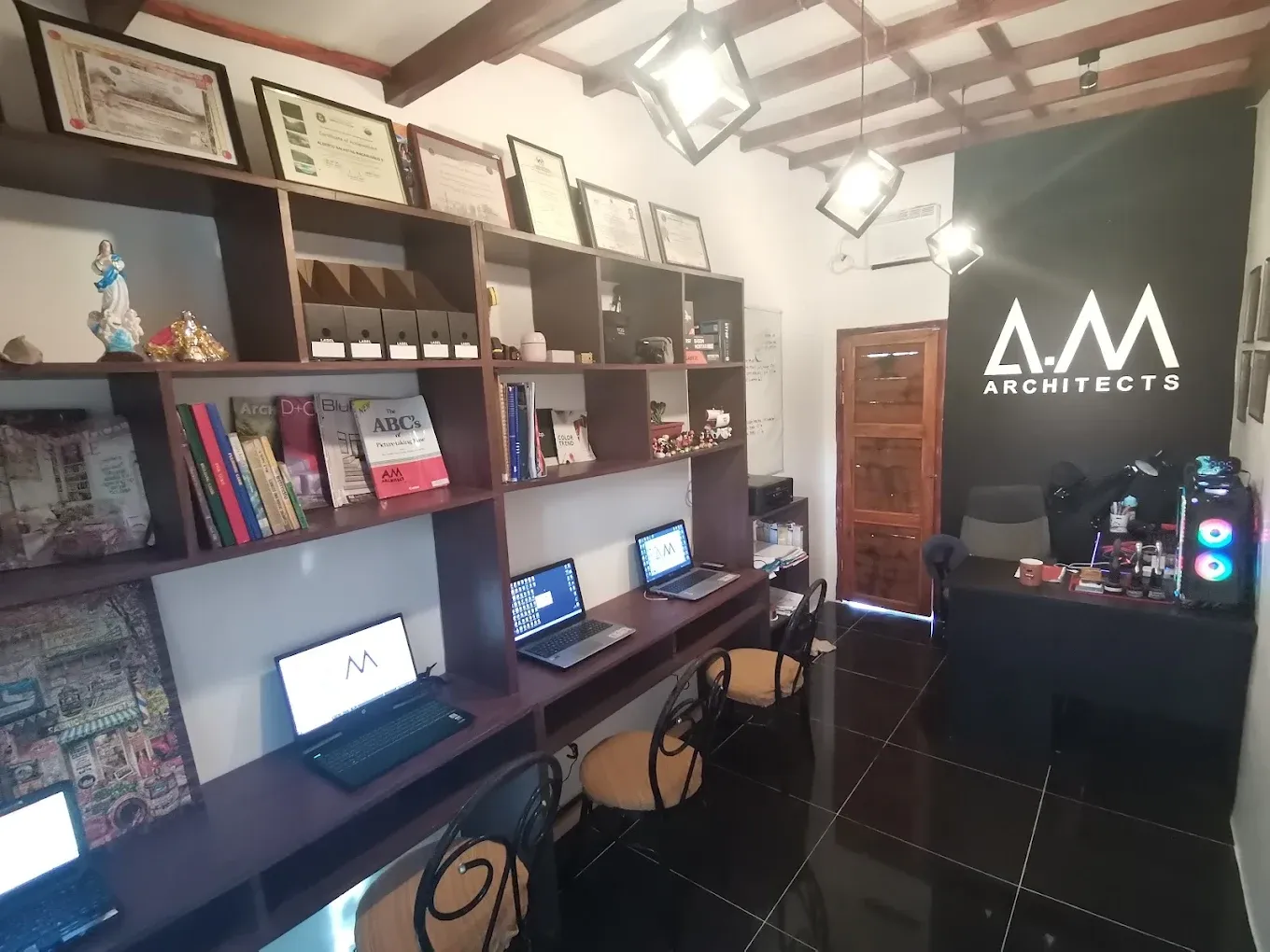 Inside the A.M Architects+ studio: shelves of books and framed licences, laptops on a long desk and the A.M Architects logo on a black wall